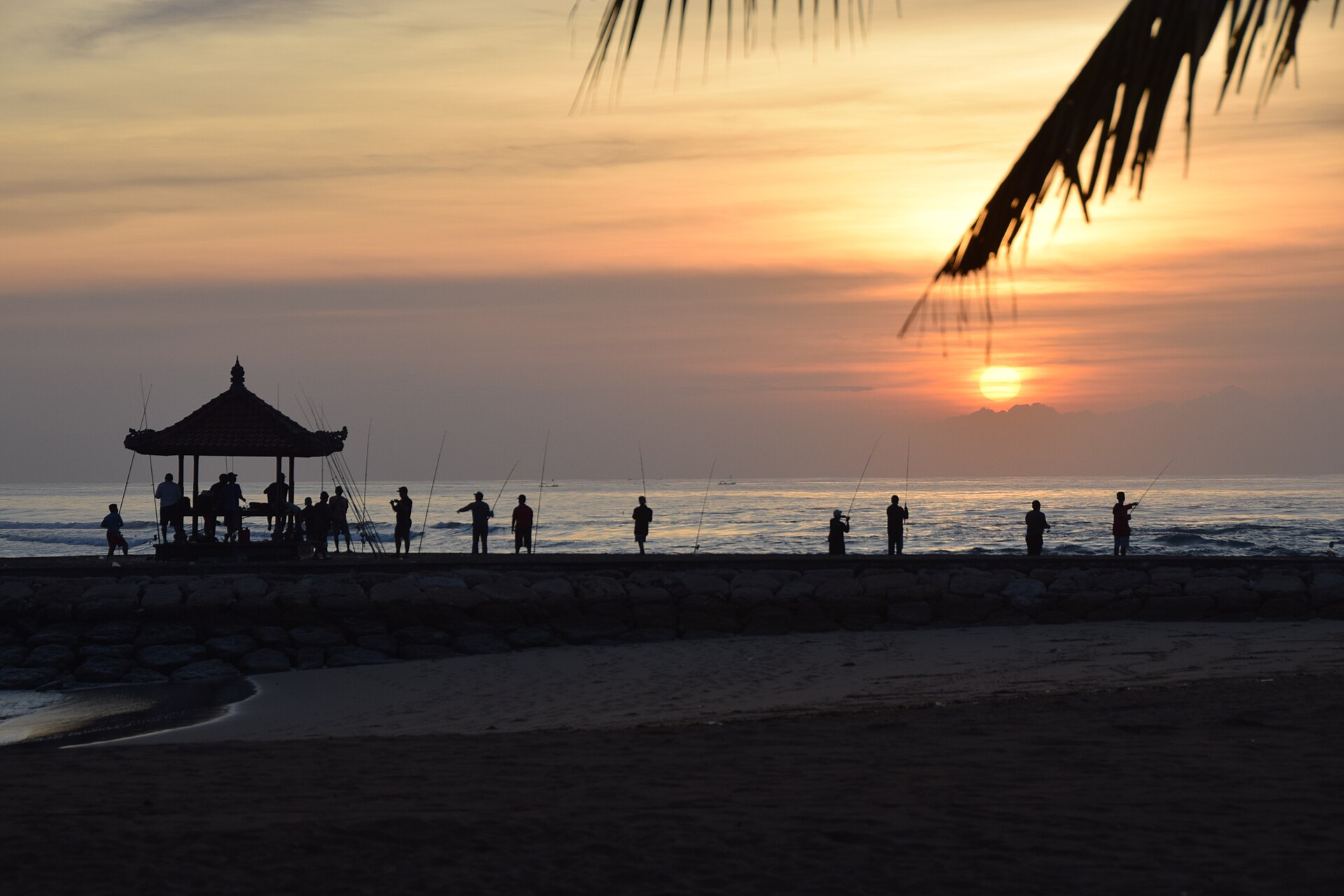 Matahari turun di Pantai Nusa Dua, dengan pemancing di atas pemecah gelombang